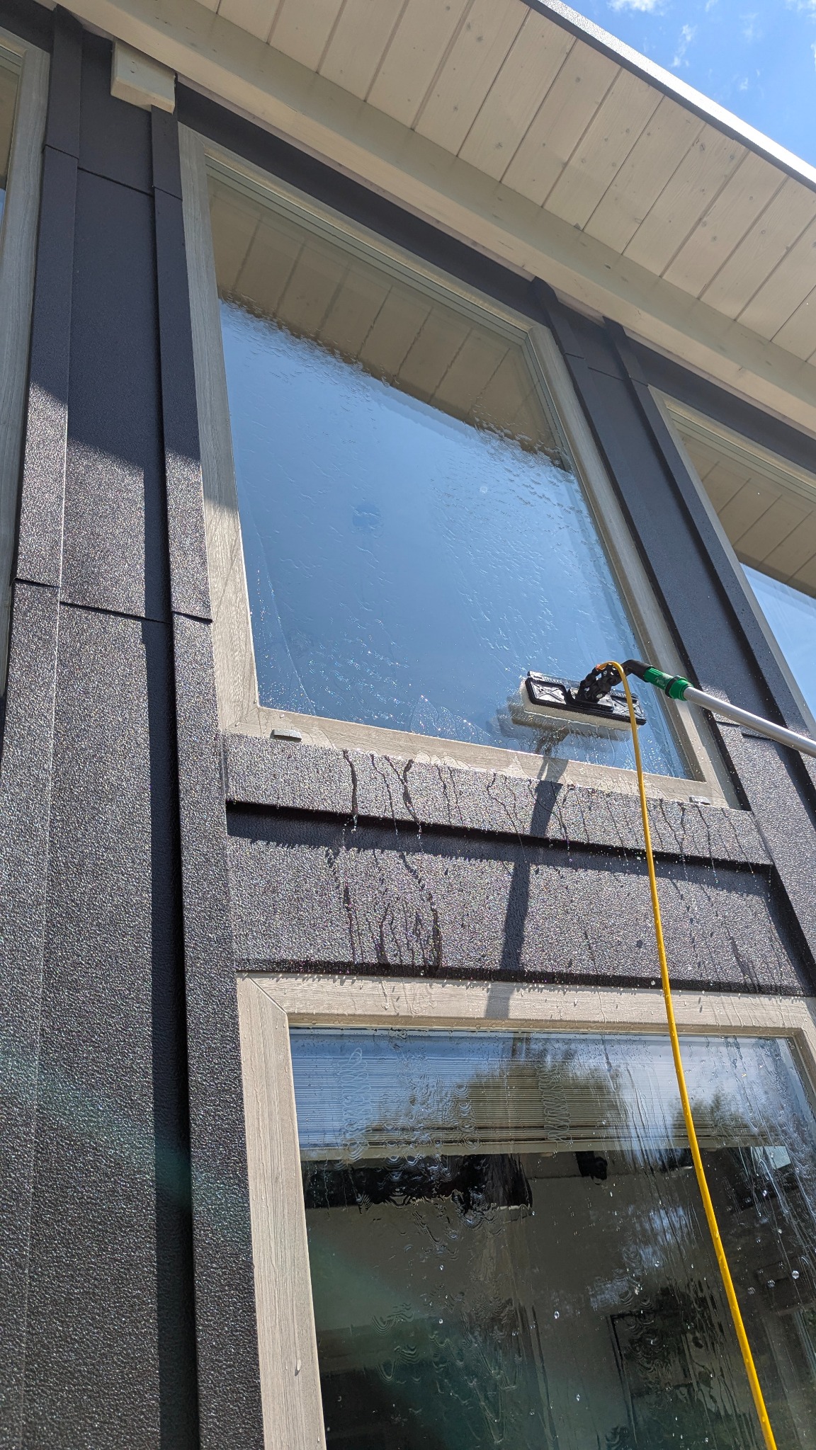 Mycie okna z zewnątrz za pomocą szczotki na teleskopowym kiju. Woda spływa po szybie. Widoczne elementy elewacji budynku i niebo.