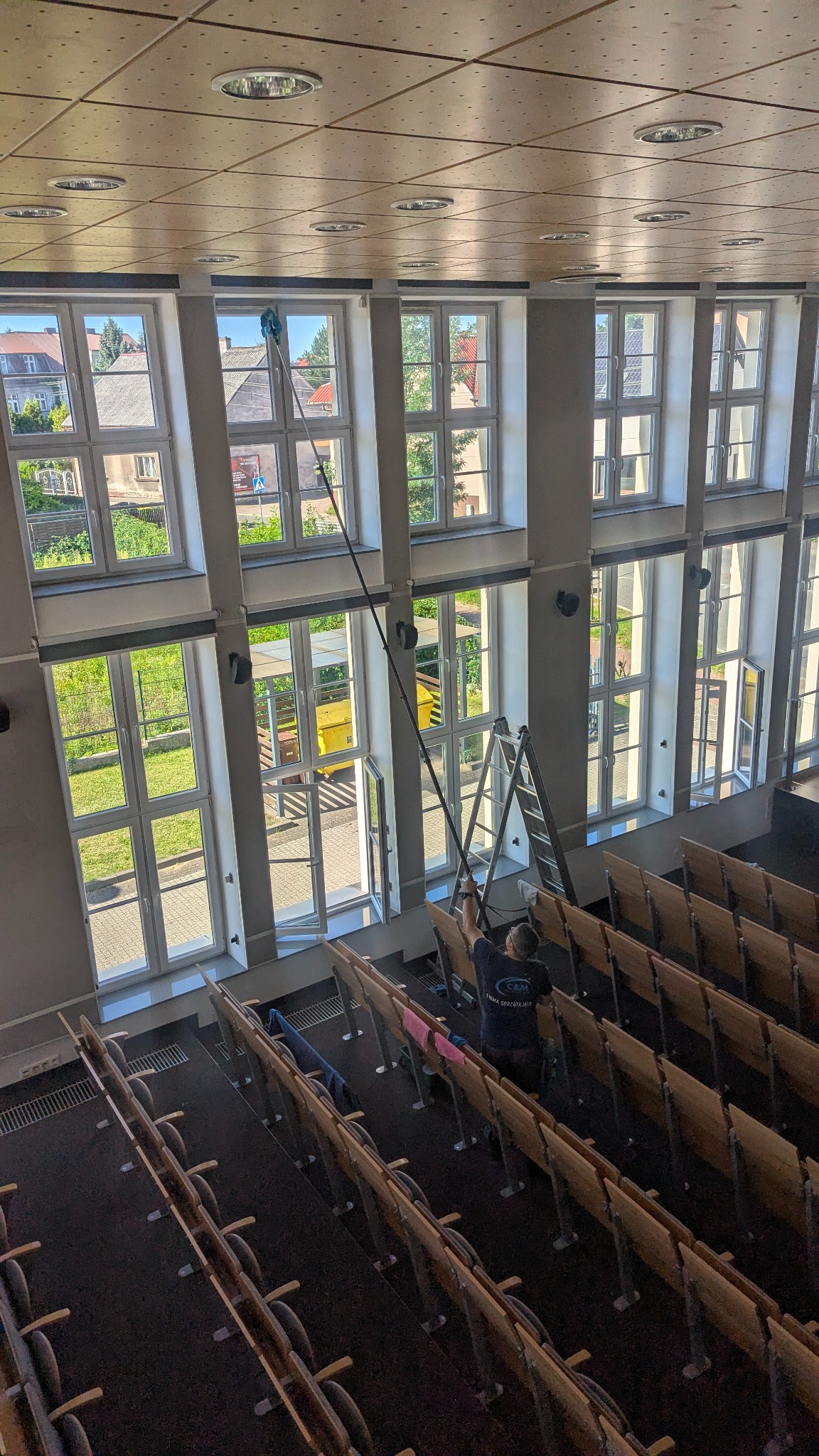 Pracownik myje wysokie okna w auli za pomocą teleskopowego kija z mopem. Aluminiowa drabina stoi obok. Widok z góry na rzędy krzeseł.