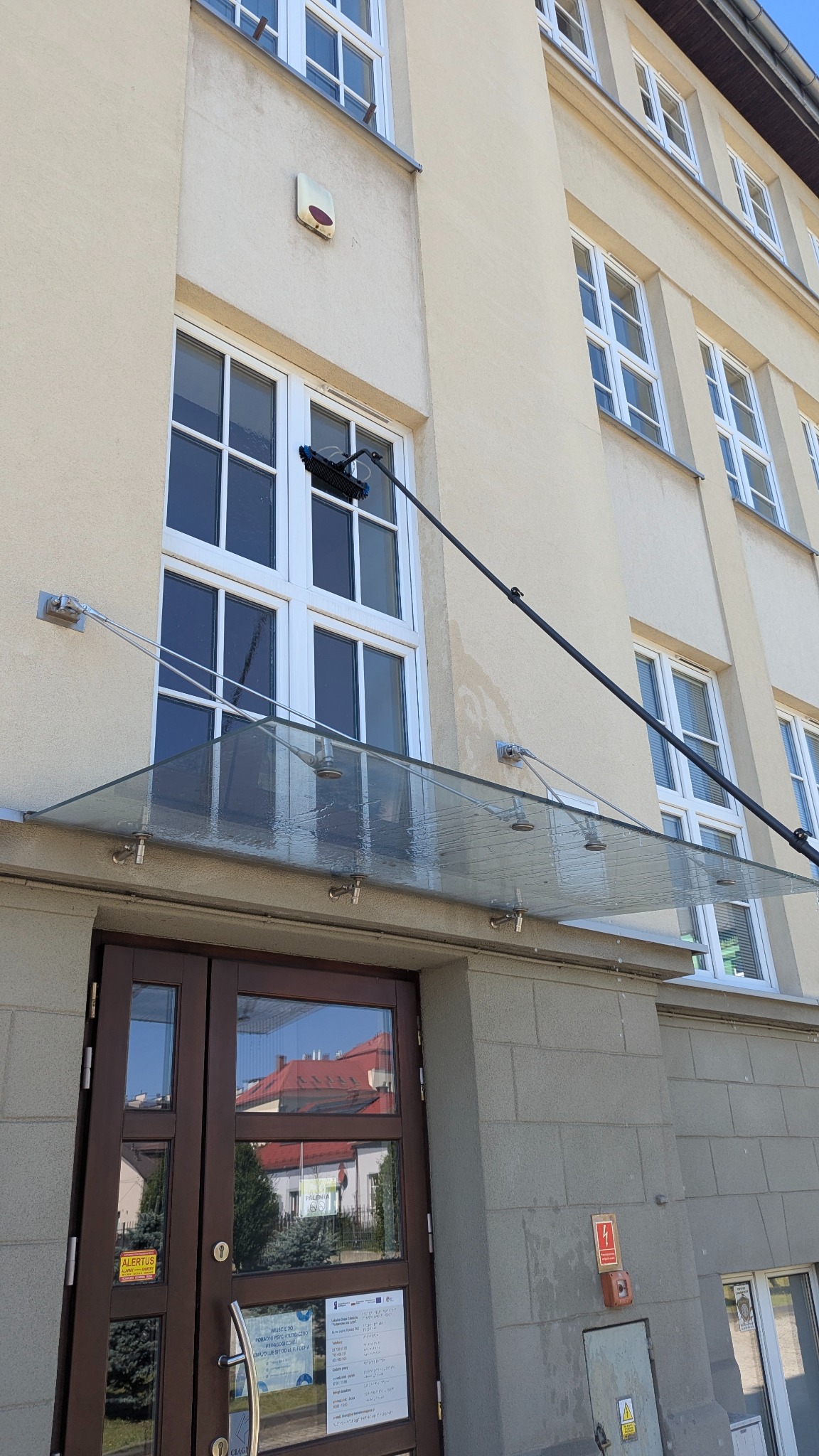 Mycie okien na wysokości w budynku z beżową elewacją. Widoczna lanca z szczotką i ślady wody po myciu okna. Ujęcie z dołu, na pierwszym planie przeszklone zadaszenie nad wejściem.