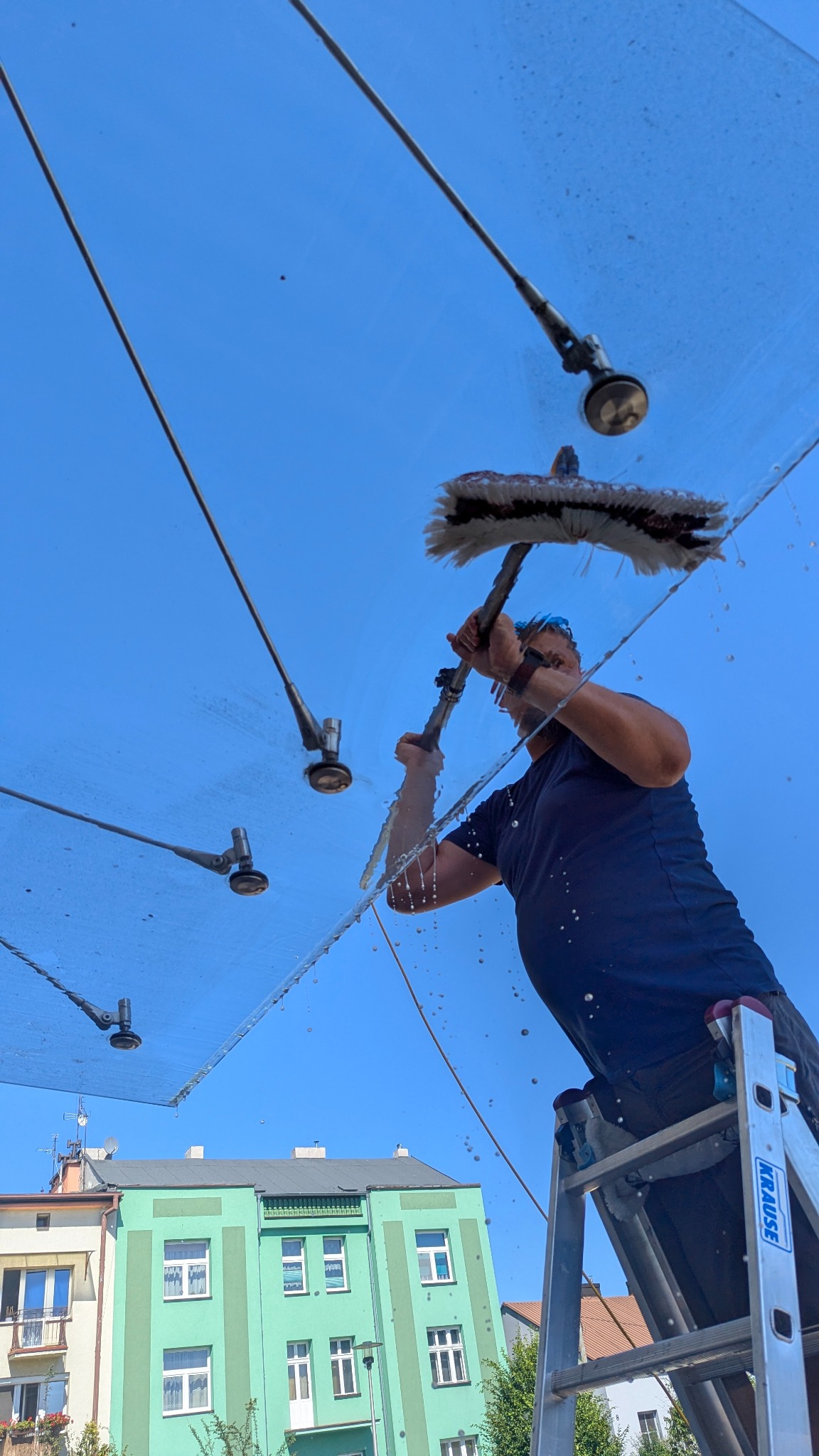 Mężczyzna na drabinie myje szklany daszek z metalowymi elementami mocującymi, w tle błękitne niebo i budynek. Widok z dołu, krople wody w powietrzu.