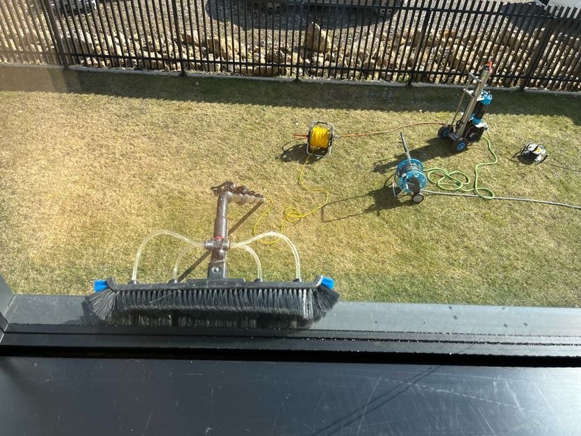 Szczotka do mycia okien z podajnikiem wody oparta o ramę okienną. W tle trawnik z rozłożonymi wężami i urządzeniem do mycia. Widok z góry, słoneczny dzień.