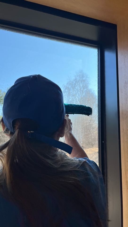 Osoba w czapce myje okno za pomocą ściągaczki, widok zza pleców, na zewnątrz widoczne drzewa.