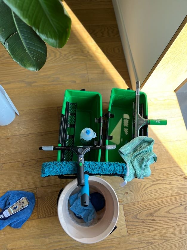 Zestaw do mycia okien: wiadro z wodą, ściągaczki, myjki i zielone pojemniki na drewnianej podłodze w jasnym świetle słonecznym, sugerujący gotowość do pracy.