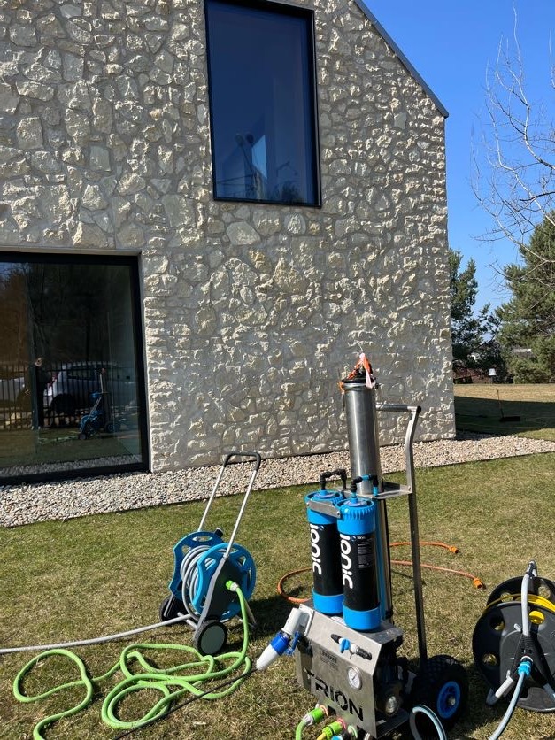 Sprzęt do mycia okien na tle domu z kamienia. Na trawniku rozłożone węże i urządzenia filtrujące wodę. Widoczne okna budynku.