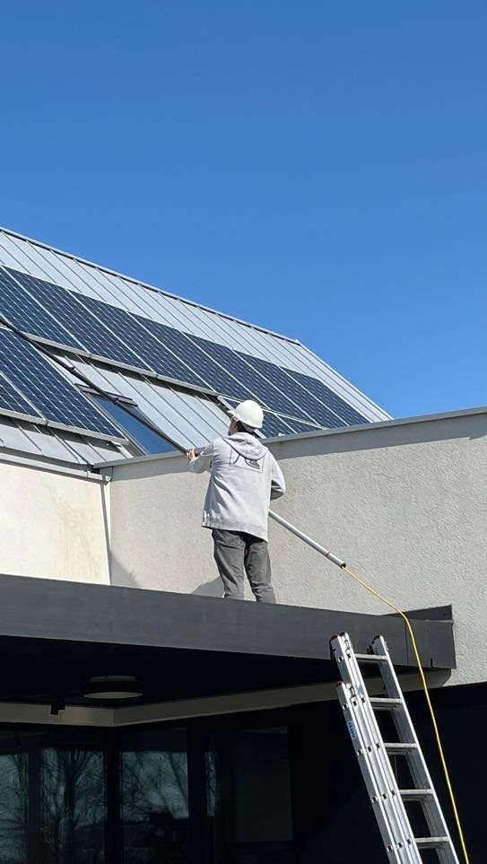 Pracownik w kasku czyści panele fotowoltaiczne na dachu budynku za pomocą długiej szczotki. Widoczna drabina i błękitne niebo.