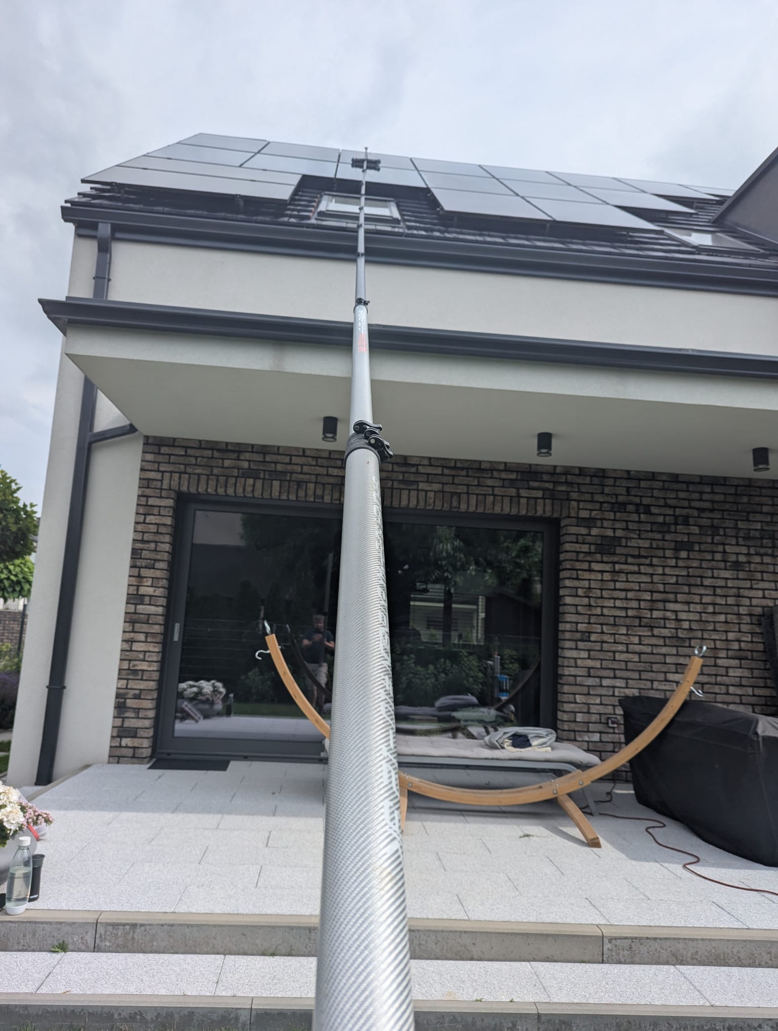 Czyszczenie paneli słonecznych na dachu domu jednorodzinnego przy użyciu teleskopowej szczotki. Widok z dołu na fasadę i panele w pochmurny dzień.