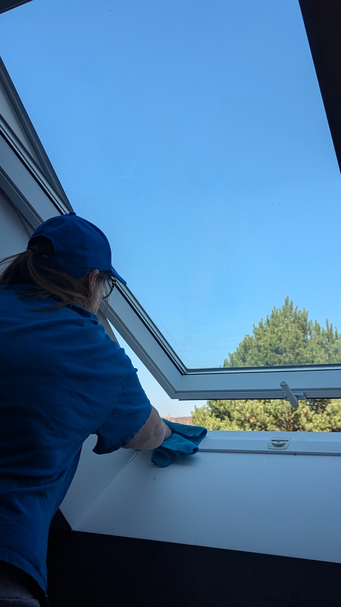 Osoba w niebieskiej koszulce czyści okno dachowe szmatką w Trzebini. Widok na błękitne niebo i drzewo. Skupienie na detalu czyszczenia ramy okiennej.