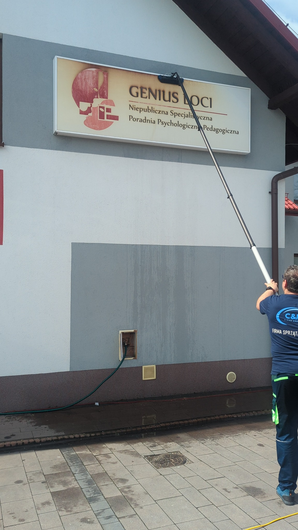 Pracownik myje elewację budynku z tablicą informacyjną 'Genius Loci' za pomocą długiego kija z myjką. Widoczny wąż doprowadzający wodę i mokra elewacja.