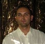 Mężczyzna w białej koszuli stojący na tle roślinności, prawdopodobnie w trakcie rozmowy lub krótkiej przerwy w pracy.