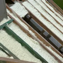 Poddasze z drewnianą konstrukcją krokwiową wypełnione białą pianą izolacyjną, widok z góry na ukośną powierzchnię, w tle zielony krajobraz.