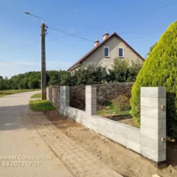 Wznoszenie ogrodzenia z szarej kostki betonowej, widoczne słupki i przęsła, w tle dom z jasnej cegły i zielony krzew, obok fragment starego kamiennego ogrodzenia.