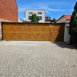 Szeroka, drewniana brama garażowa z trzema segmentami, zabezpieczona zamkami, na tle brukowanej nawierzchni podjazdu, z widocznym fragmentem kamiennego muru i budynków w tle.