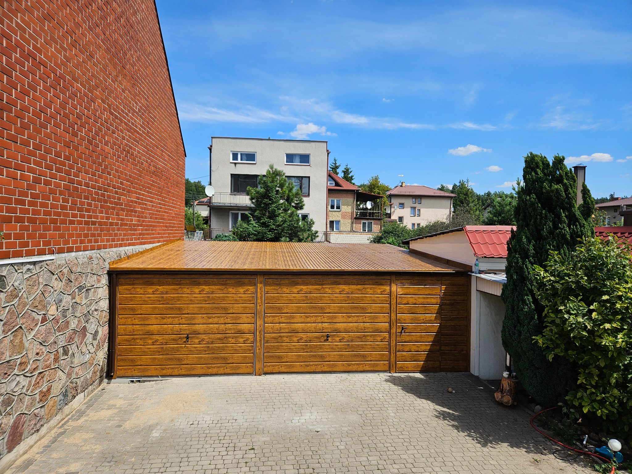 Trzysegmentowy garaż blaszany z drewnopodobną fakturą, przylegający do ceglanego budynku, z widocznym brukowanym podjazdem i fragmentami sąsiednich budynków w tle pod błękitnym niebem.