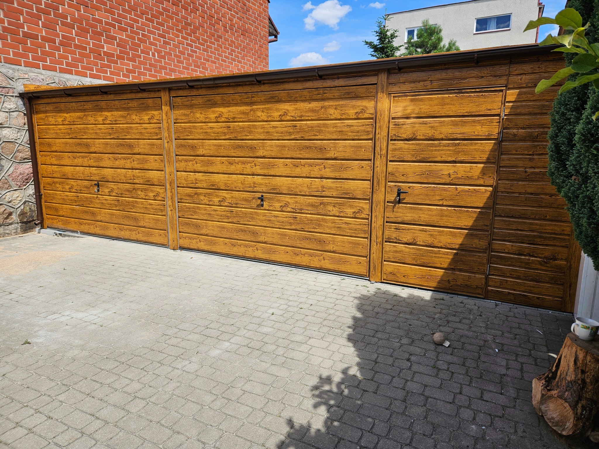 Zewnętrzny, modułowy garaż z blachy imitującej drewno, z trzema segmentami bram i dodatkowymi drzwiami wejściowymi, na tle brukowanej nawierzchni i fragmentu budynku z czerwonej cegły.