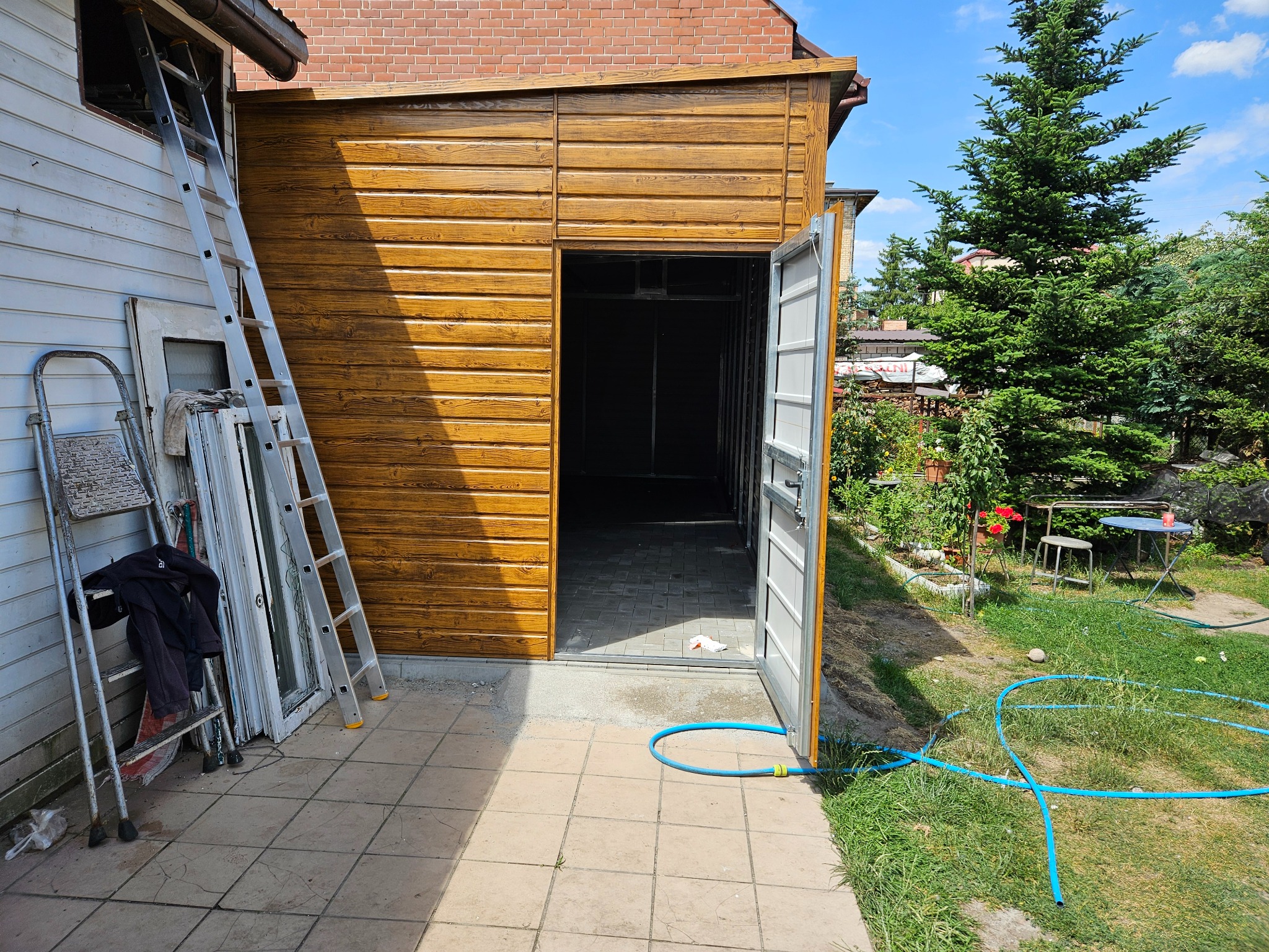 Otwarty, brązowy garaż blaszany z widocznymi drzwiami i wnętrzem, na zewnątrz drabina oparta o ścianę budynku, obok złożone okna PCV oraz niebieski wąż ogrodowy na trawie