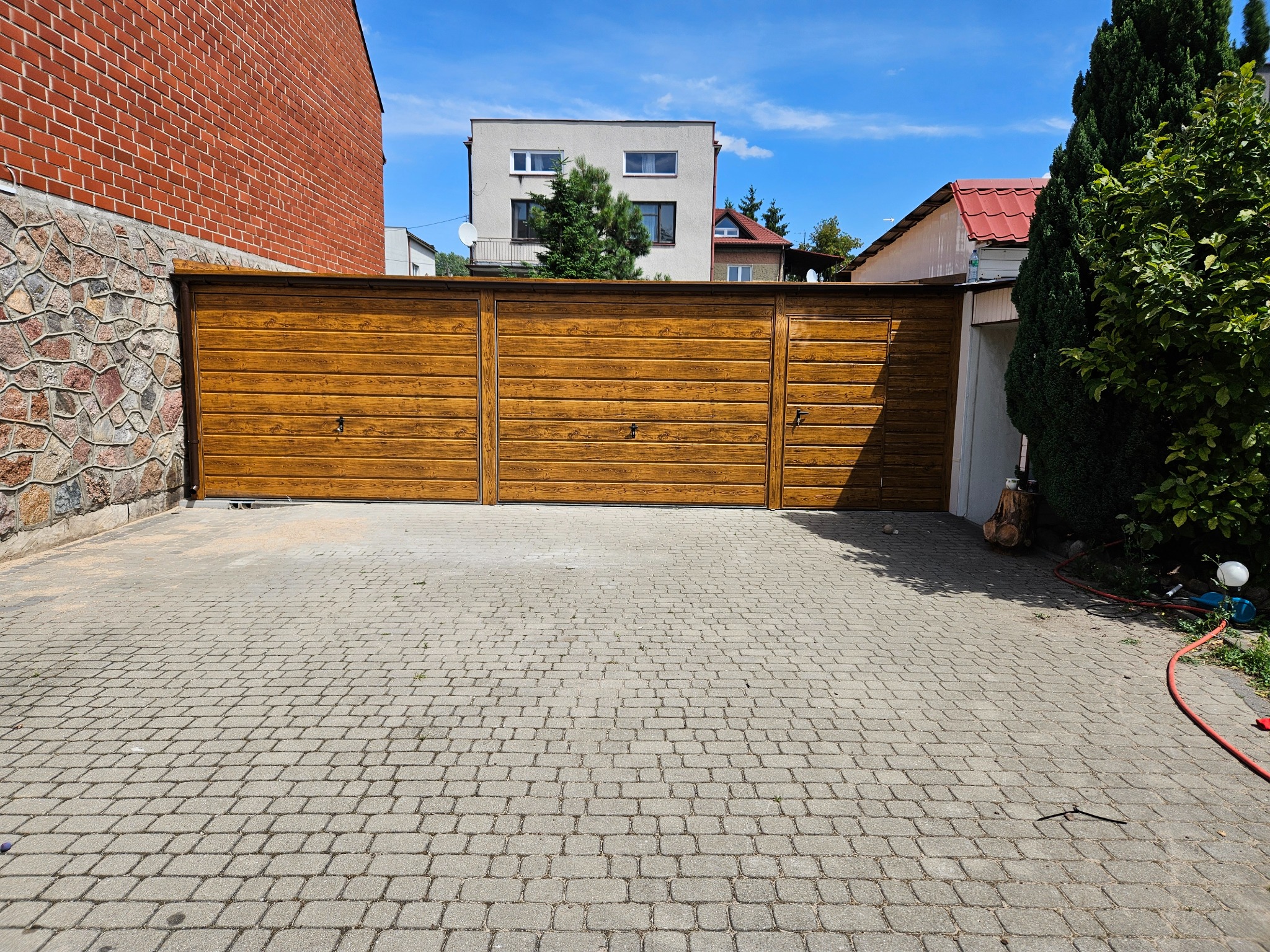 Szeroka, drewniana brama garażowa z trzema segmentami, zabezpieczona zamkami, na tle brukowanej nawierzchni podjazdu, z widocznym fragmentem kamiennego muru i budynków w tle.