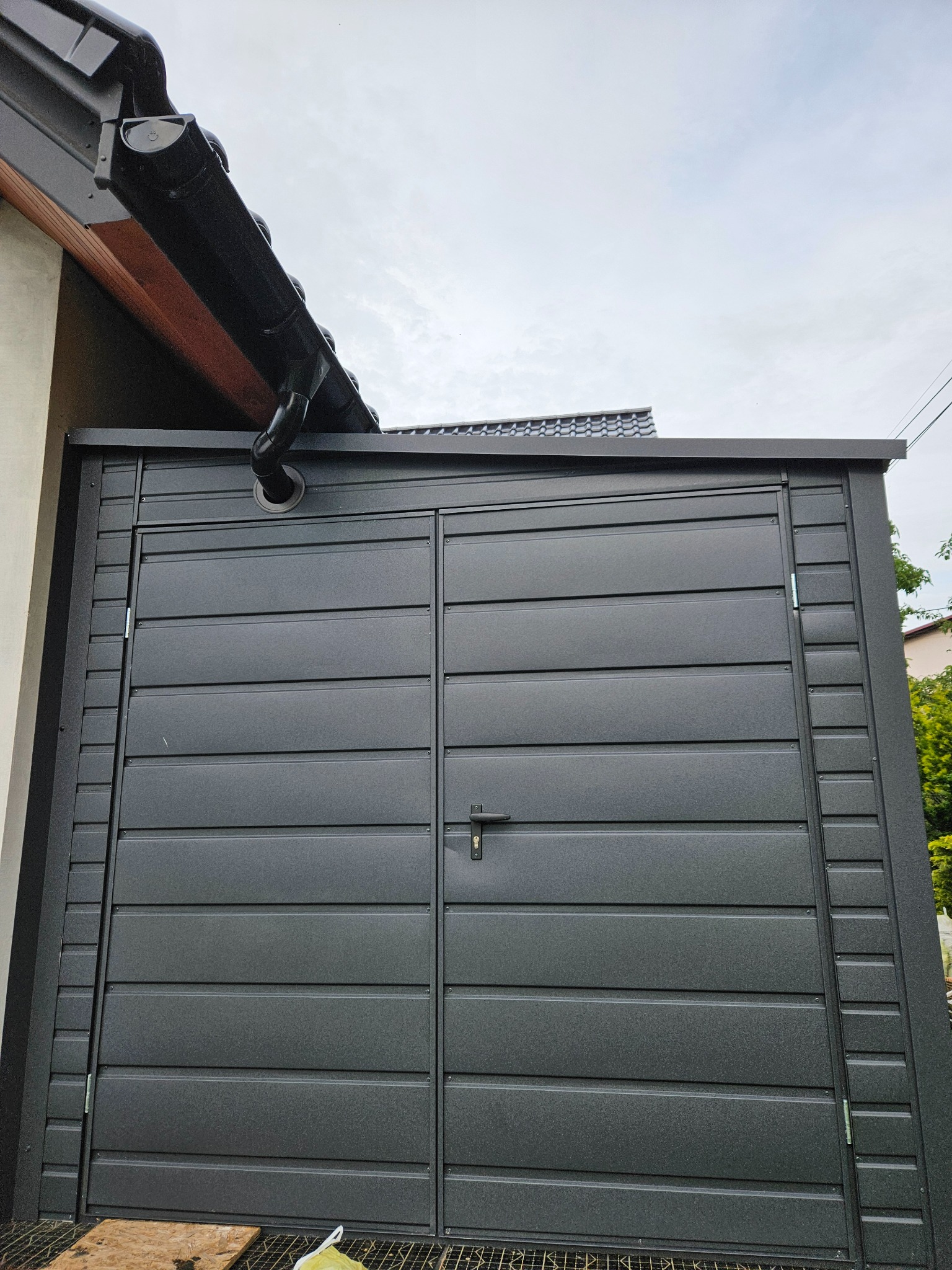 Nowoczesny, grafitowy garaż blaszany z dwuskrzydłowymi drzwiami, widoczny fragment orynnowania domu w tle, ujęcie od dołu.
