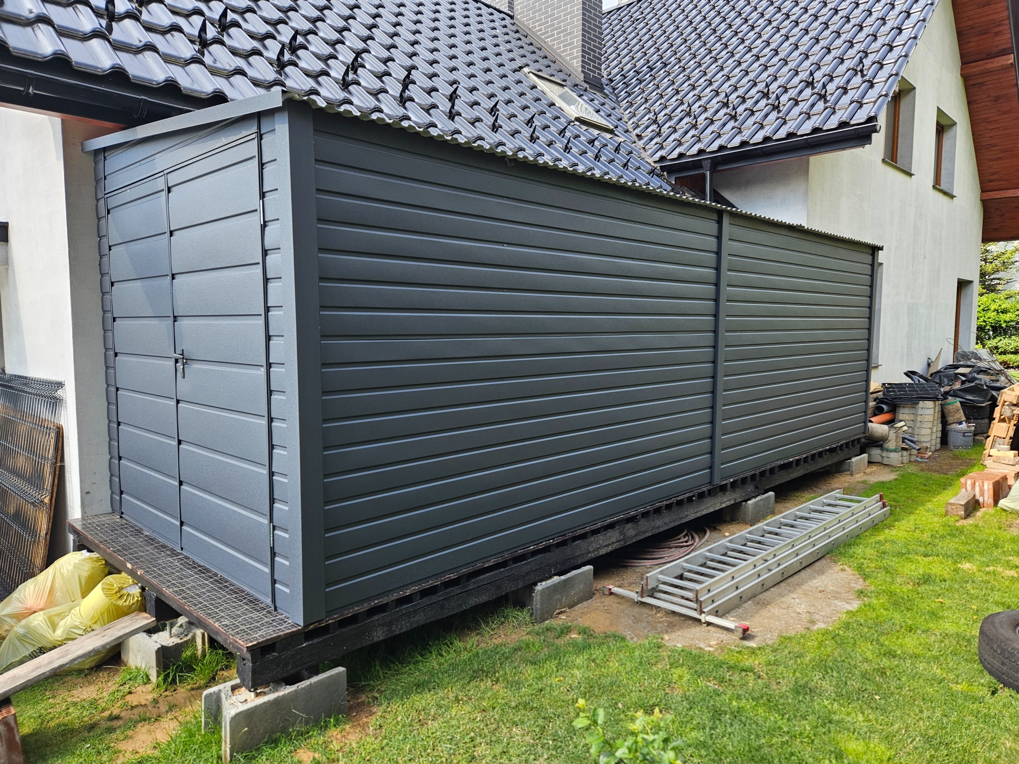 Szary garaż blaszany przylegający do domu z czarnym dachem, widoczna drabina i materiały budowlane na trawniku.