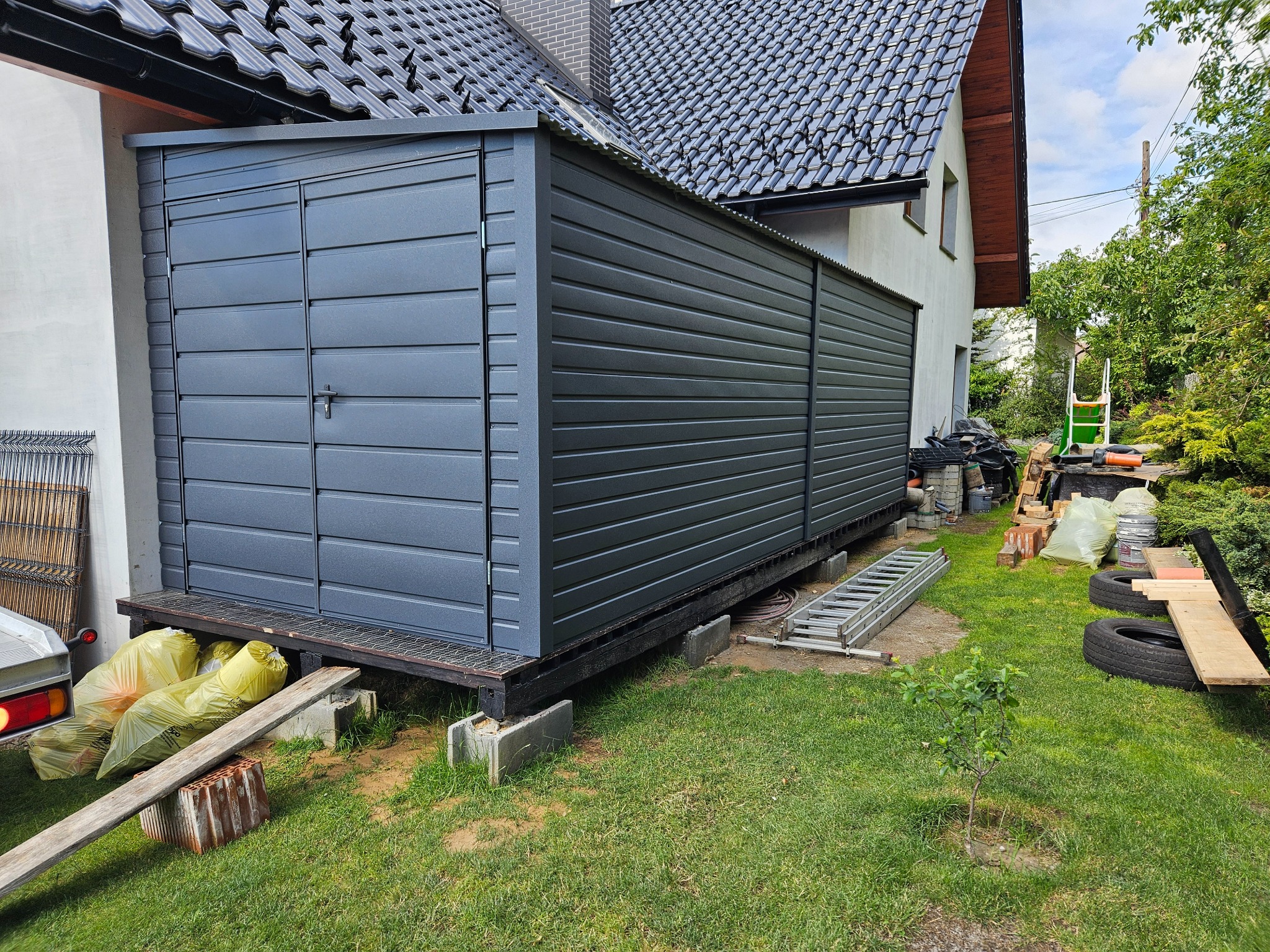 Szary garaż blaszany z dwuskrzydłowymi drzwiami, umieszczony na podwyższeniu z betonowych bloczków, widok z boku, w tle dom z ciemną dachówką.