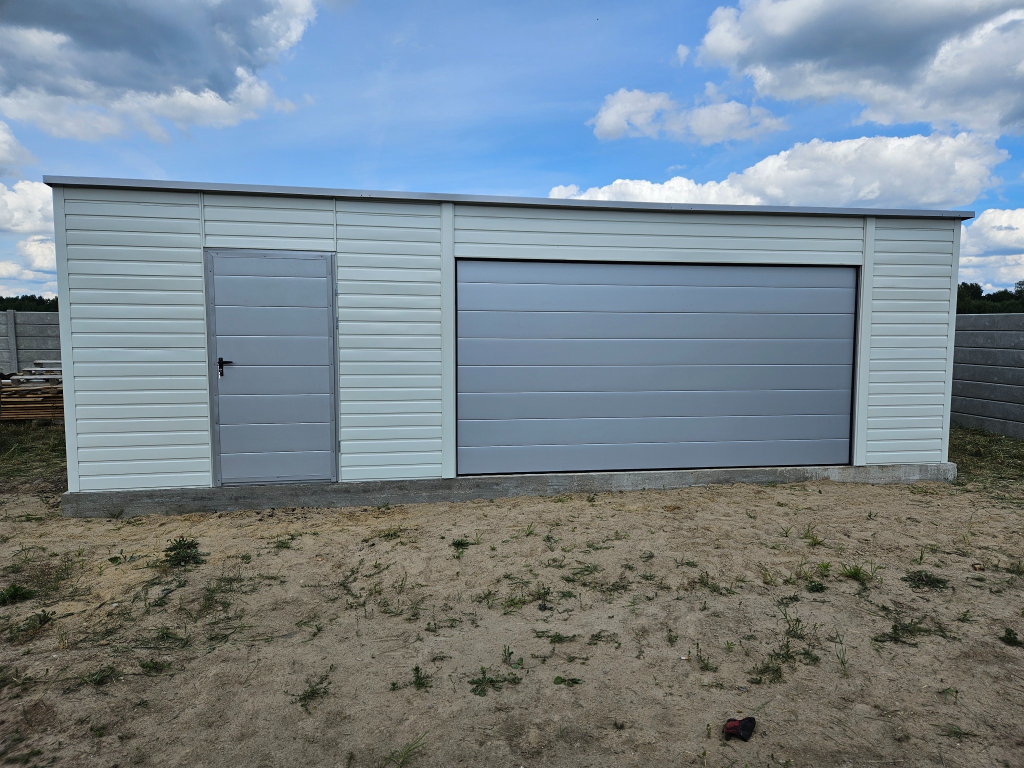 Gotowy garaż blaszany z dwoma wejściami: bocznymi drzwiami i bramą rolowaną, na nierównym, piaszczystym terenie z widocznymi chmurami na niebie.