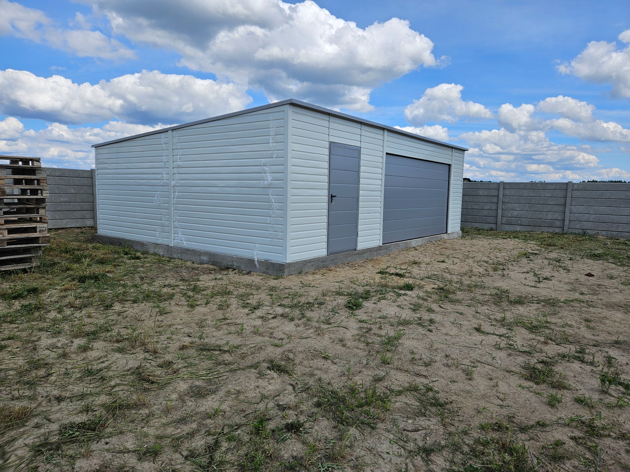 Gotowy garaż blaszany z bramą segmentową i drzwiami w Raciechowicach, widoczny na tle betonowego ogrodzenia i błękitnego nieba z chmurami.