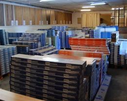 Magazyn z zapasem paneli podłogowych w paczkach, widoczne różne wzory i kolory, tło stanowią drewniane deski ułożone pionowo.