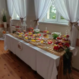 St&oacute;ł z białym obrusem i złotym bieżnikiem, zastawiony paterami z ciastami i owocami, dekorowany kwiatami i lampionem, okno z białymi zasłonami w tle.