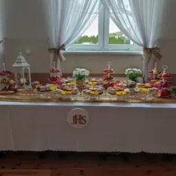 Stół z białym obrusem, złotym bieżnikiem i dekoracją IHS, zastawiony owocami i ciastami na szklanych paterach, białe kwiaty w wazonach, lampion ze świecą, okno z białymi firankami w tle.