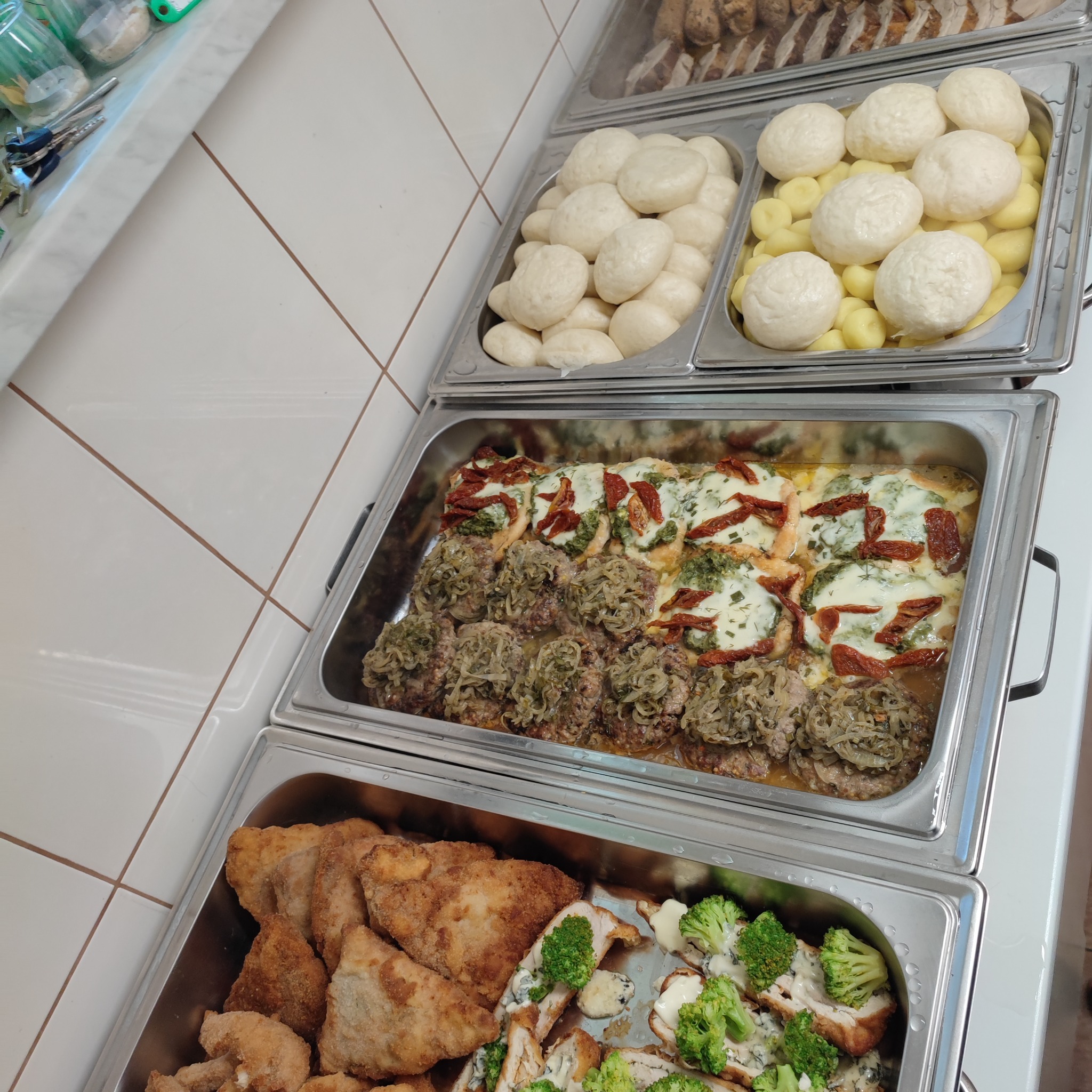 Bufet cateringowy z różnorodnymi daniami: kluski na parze obok ziemniaków, kotlety mielone z duszoną cebulą, kurczak z suszonymi pomidorami i pesto, oraz kotlety schabowe z brokułami w sosie serowym.