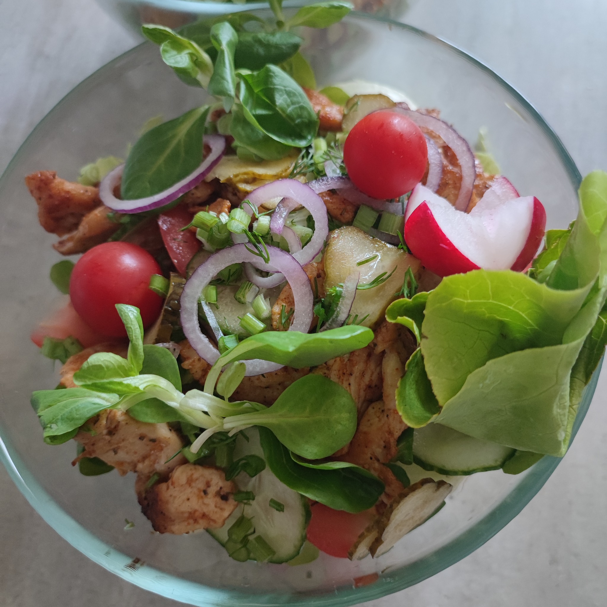Szklana misa z sałatką: grillowany kurczak, sałata, pomidorki cherry, czerwona cebula, ogórek kiszony, rzodkiewka i szczypiorek, całość skropiona sosem.