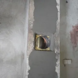 Montaż przewodu wentylacyjnego w ścianie, widoczne świeże obłożenie zaprawą wokół otworu, fragment instalacji elektrycznej w tle.