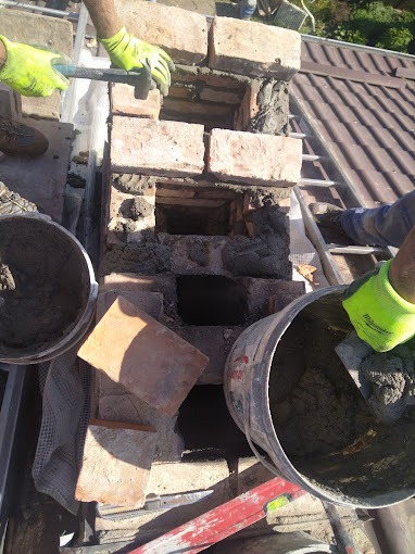 Prace murarskie na dachu: wznoszenie komina z cegły, widoczne zaprawa, cegły, drabina dachowa i dłonie w roboczych rękawicach. Częściowo zrekonstruowany komin z otworami.