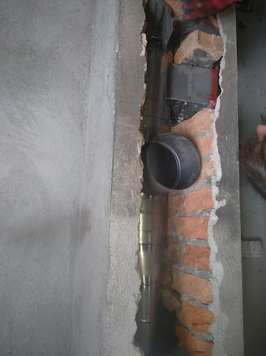 Montaż metalowego przewodu spalinowego w kominie z cegły, widoczne fragmenty ściany z betonu i cegieł, zarys dłoni w rękawicy ochronnej w górnym rogu.