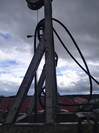 Metalowa konstrukcja z kołem zębatym i przewodami na tle pochmurnego nieba i zabudowań z czerwonymi dachami, widok z dołu.