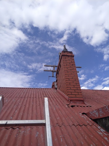 Ceglany komin z anteną na dachu z blachy falistej, widoczna drabina przystawiona do dachu na tle błękitnego nieba z chmurami.