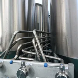 Instalacja przemysłowa z dużymi, metalowymi zbiornikami i skomplikowaną siecią rur ze stali nierdzewnej, zawory z niebieskimi uchwytami, etykieta 'PASTERYZATOR'.