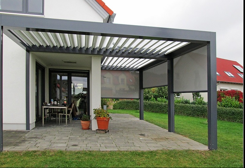 Nowoczesna, grafitowa pergola z regulowanymi lamelami i roletami przeciwsłonecznymi, przylegająca do białego domu z ceglanym dachem, na brukowanym patio z roślinami w donicach i widokiem na zadbany...