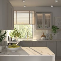 Pracownia Architektury Wnętrz Klaudia Woźniak - Jasna kuchnia z marmurowym blatem, wyposażona w szare szafki z delikatnymi złotymi uchwytami, przeszklone szafki wiszące, złota armatura i widok na zieleń za oknem, uzupełniona roślinami w wazonach...
