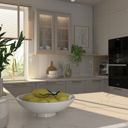 Pracownia Architektury Wnętrz Klaudia Woźniak - Jasna kuchnia z marmurowym blatem, na którym stoi biała misa z cytrynami i wazon z zieloną rośliną. W tle okno z widokiem na zieleń, zabudowane szafki z przeszklonymi drzwiczkami oraz piekarnik...