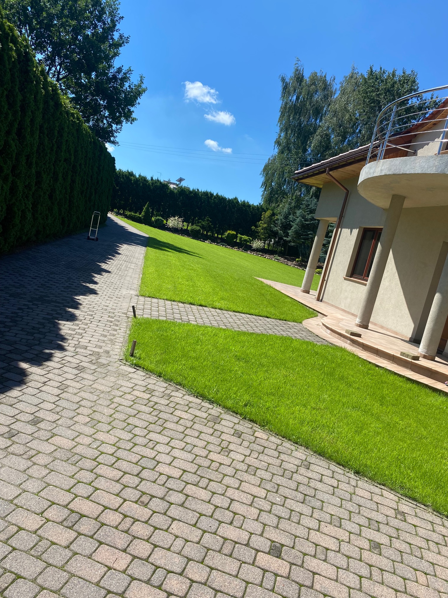 Ułożona kostka brukowa prowadząca do domu z zadbanym trawnikiem i wysokim żywopłotem w słoneczny dzień.