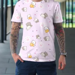 Mężczyzna prezentuje t-shirt z nadrukiem pastelowych kotów, łapek i geometrycznych wzorów na jasnoróżowym tle, widoczne tatuaże na ramieniu i srebrny zegarek na nadgarstku.