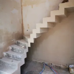 Wnętrze w trakcie remontu z widocznymi betonowymi schodami w stanie surowym, ściany częściowo otynkowane, na podłodze widoczne rury i zabrudzenia, narzędzie murarskie na jednym ze stopni.