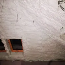 Poddasze ocieplone białą pianką PUR z widocznymi oknami dachowymi, wystającymi przewodami i metalowymi elementami mocującymi piankę, na dole ściana z zaciekami.