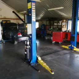 Wnętrze warsztatu samochodowego z podnośnikiem dwukolumnowym, szary samochód z otwartą maską i bagażnikiem, narzędzia i szafki w tle.