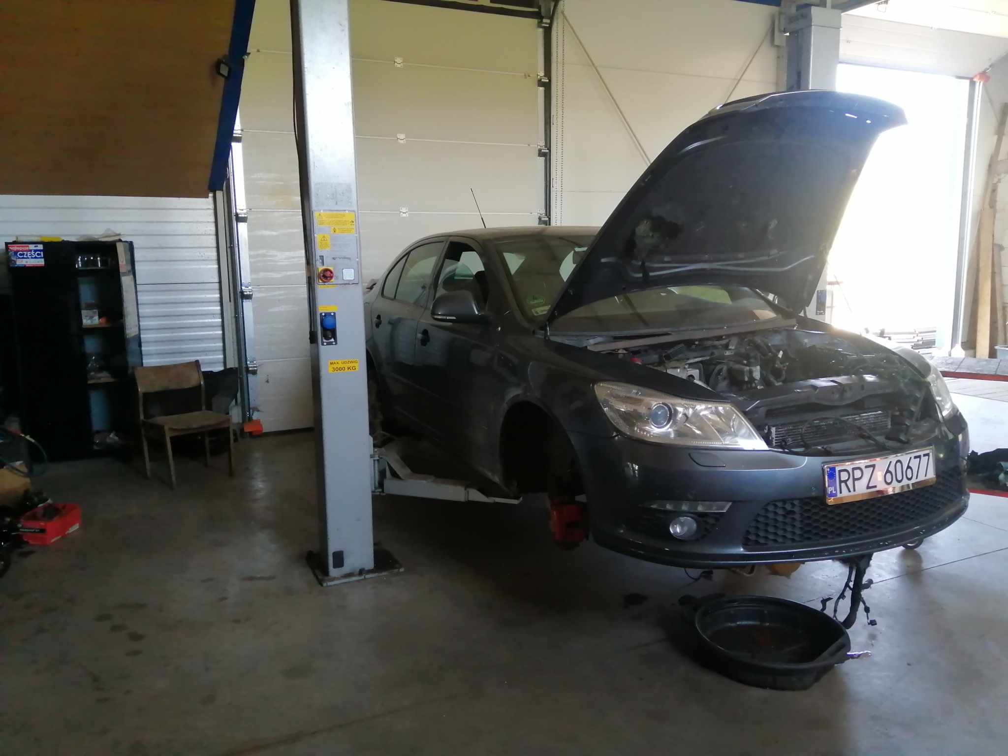 Szary samochód osobowy na podnośniku warsztatowym z otwartą maską, widoczny silnik, brak jednego koła, pod spodem miska ociekowa.