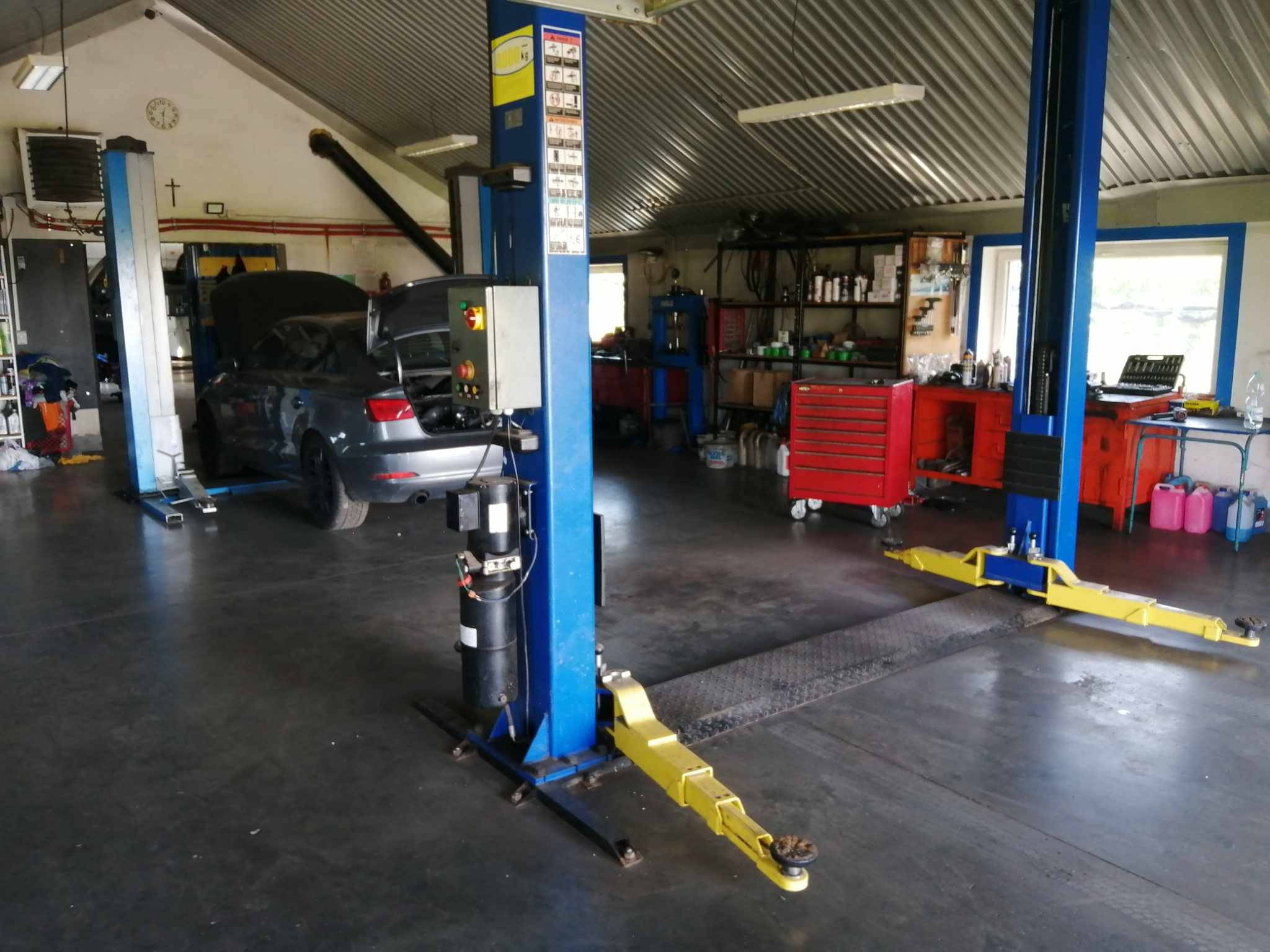 Wnętrze warsztatu samochodowego z podnośnikiem dwukolumnowym, szary samochód z otwartą maską i bagażnikiem, narzędzia i szafki w tle.