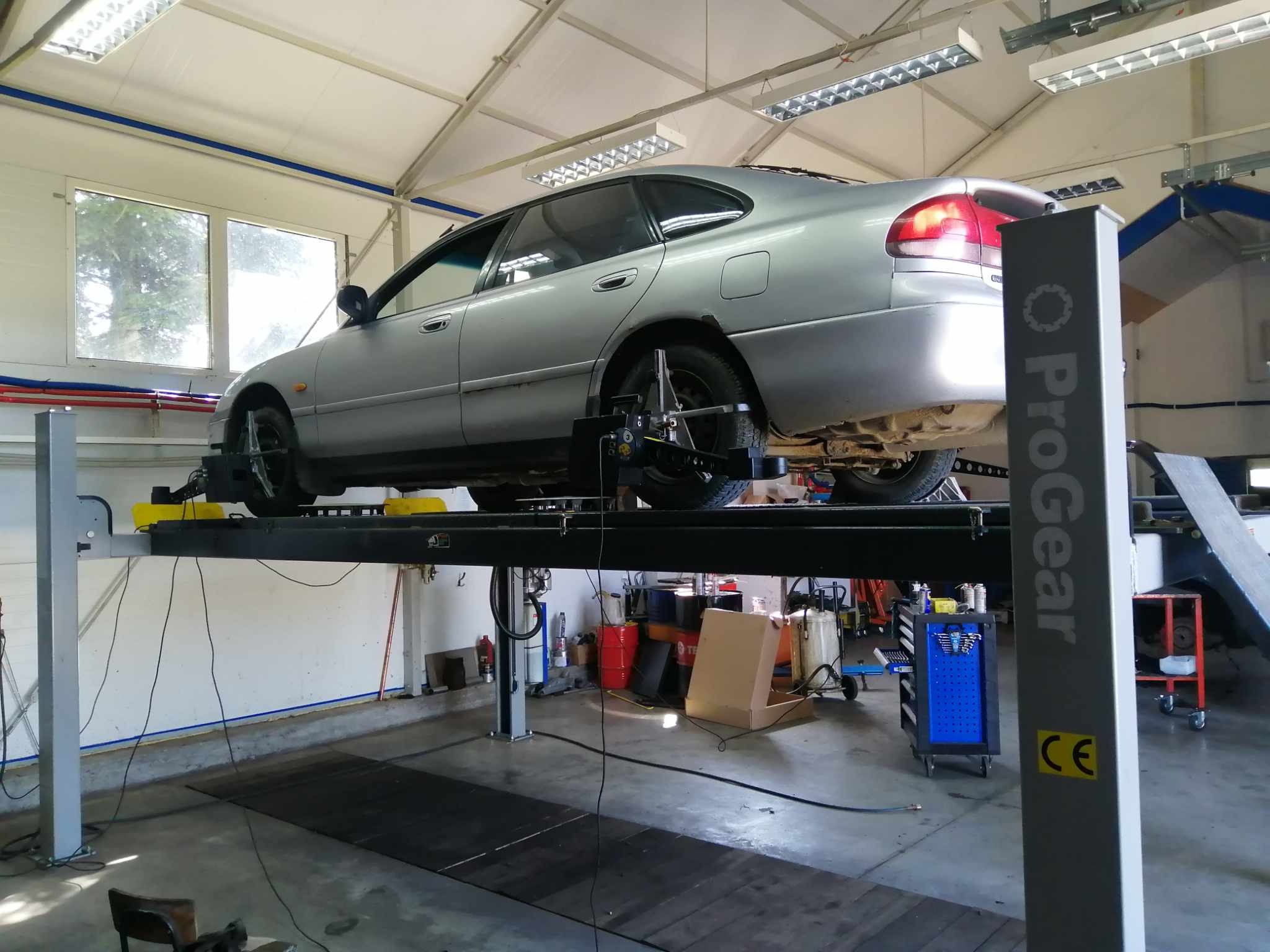 Srebrny sedan na podnośniku warsztatowym, z widocznym oprzyrządowaniem do geometrii kół, w tle okna i wyposażenie warsztatu.