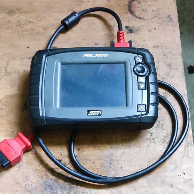 Urządzenie diagnostyczne SUN POL3000 z wyświetlaczem i przewodami zakończonymi czerwonymi wtyczkami, leżące na drewnianym blacie.