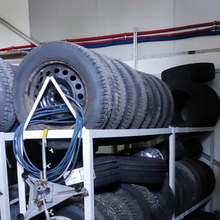 Rząd opon samochodowych na metalowym regale w warsztacie, widoczny sprzęt do wyważania kół i niebieski kabel.