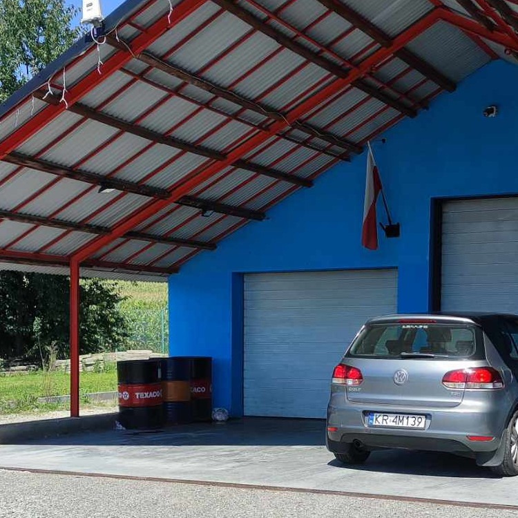 Szary Volkswagen Golf parkuje przed niebieskim warsztatem z czerwonym zadaszeniem, obok beczki Texaco i flaga Polski.
